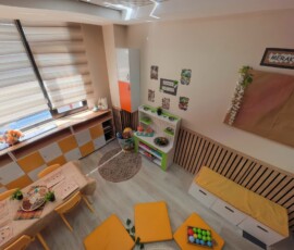 Panjurlu pencere önünde ahşap çalışma tezgâhı ve turuncu minderler